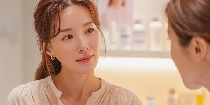 광고 모델 — 화장품 선반 앞에서 마주 앉은 사람에게 미소 짓는 여성